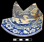 Pearlware saucer printed underglaze in blue. Design based on print entitled “Nuneham Courtenay, Bridge and Cottage” by S. Owen and published in 1811. Continuous repeating floral border of wild roses. This pattern was used by most major and many minor potteries in the second quarter of the nineteenth century (Coysh and Henrywood 1982, 1989). This design, although based on a print of an actual location, is a better fit in the Pastoral category, for its rural location and fishing theme.